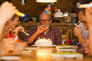 Man at head of table with friends on his birthday blowing noisemakers and a cake with lit candles